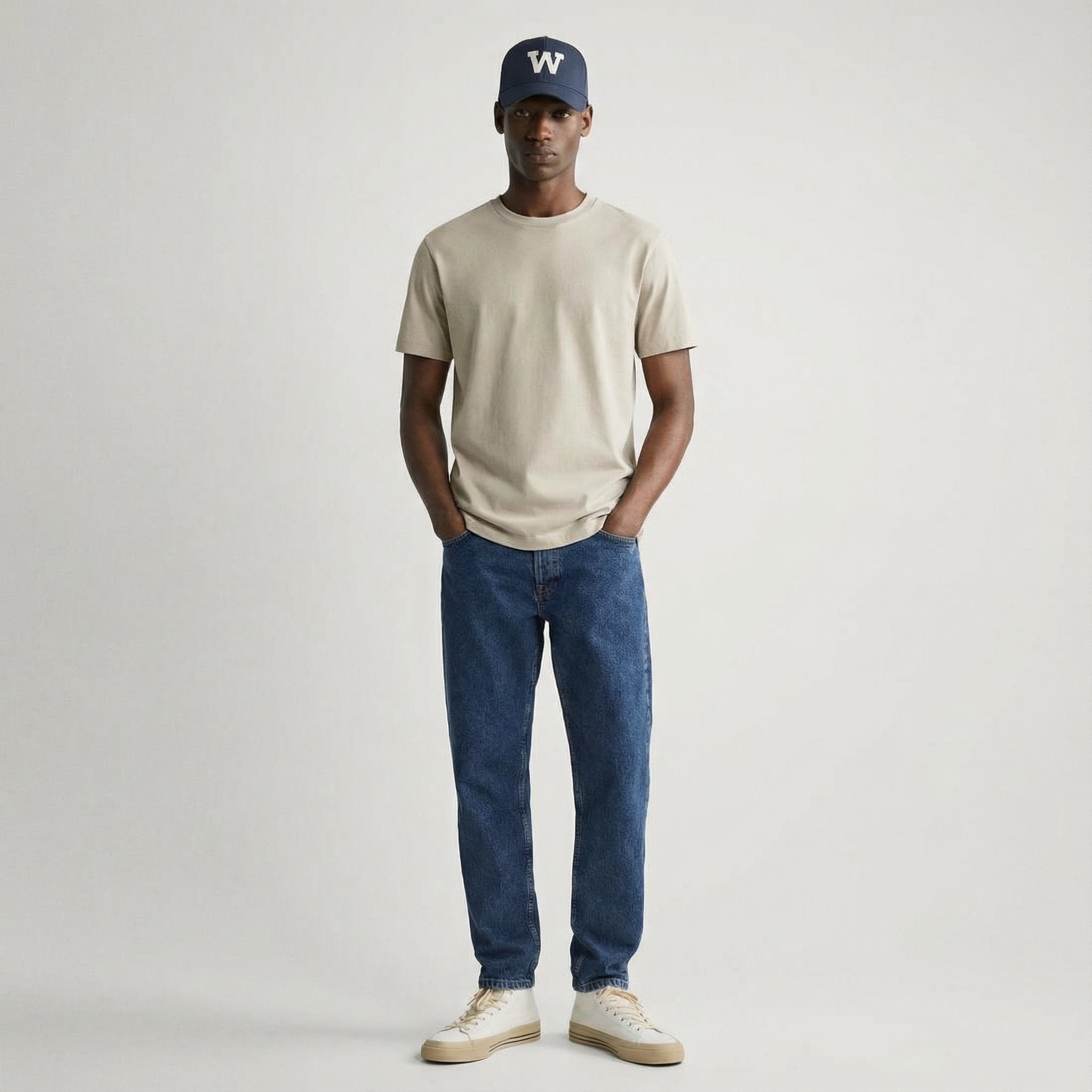 Man wearing a beige t-shirt, blue jeans, and a navy cap with a white logo on a plain background